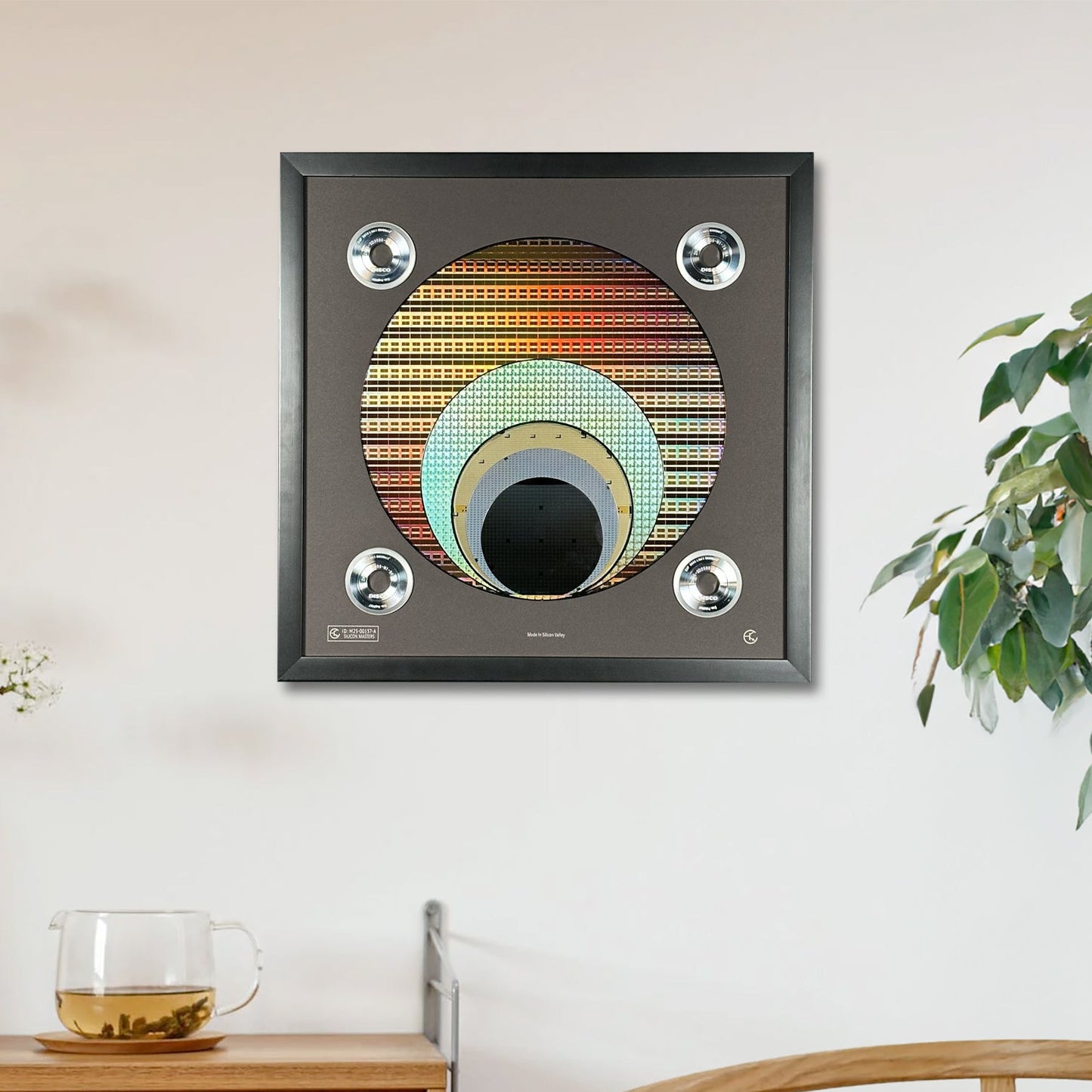 A framed square wall art piece featuring concentric silicon wafer in warm and cool tones, hung on a white wall above a wooden cabinet with a glass cup and a wooden chair nearby.