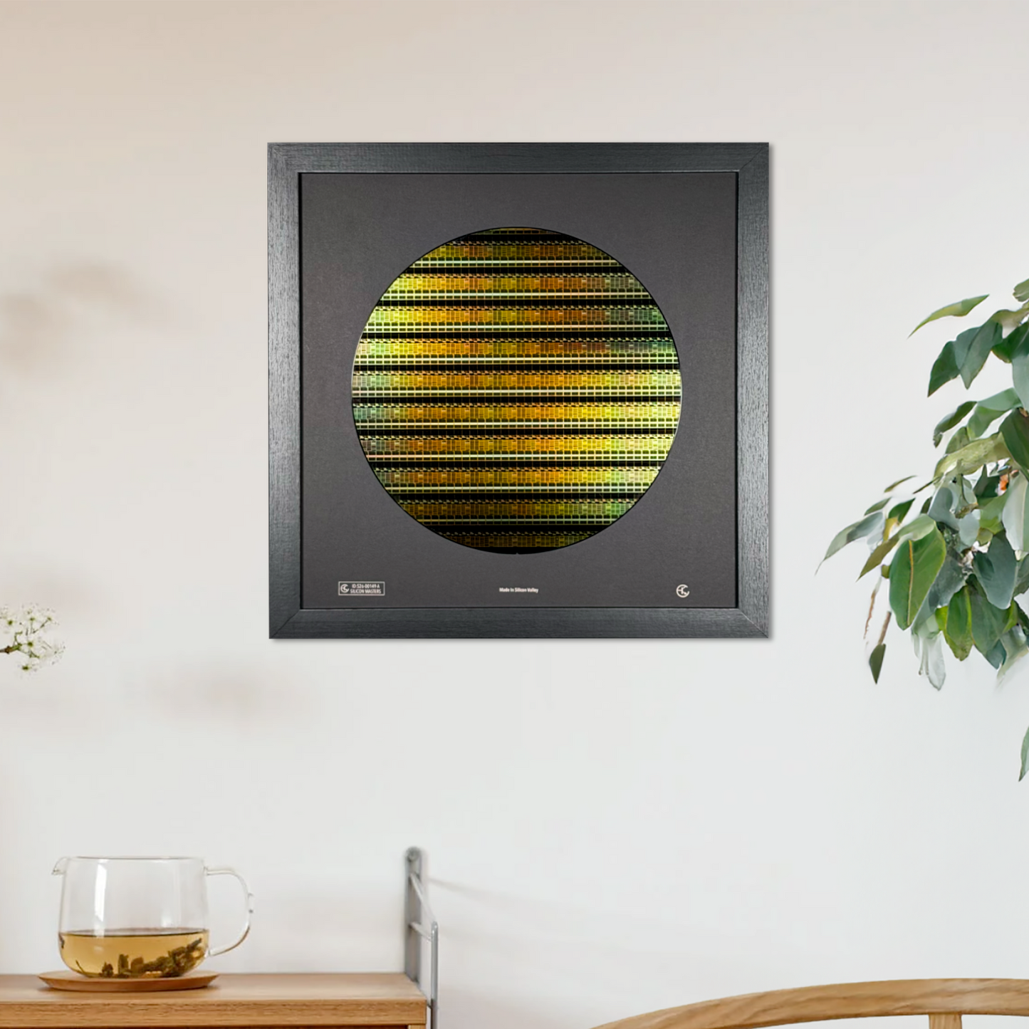 A square black-framed wall art piece hanging on a white wall, featuring a silicon wafer with horizontal yellow, green, and gold stripes. The scene includes a wooden sideboard, a glass cup, a wooden chair, and a potted plant.