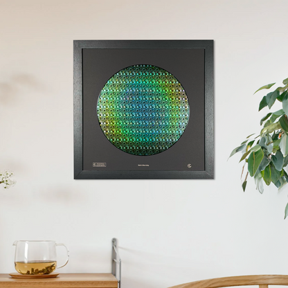 A square black-framed wall art piece featuring a silicon wafer, iridescent green and blue pattern, displayed on a white wall in a styled room setting with a wooden cabinet, chair, and potted plant.