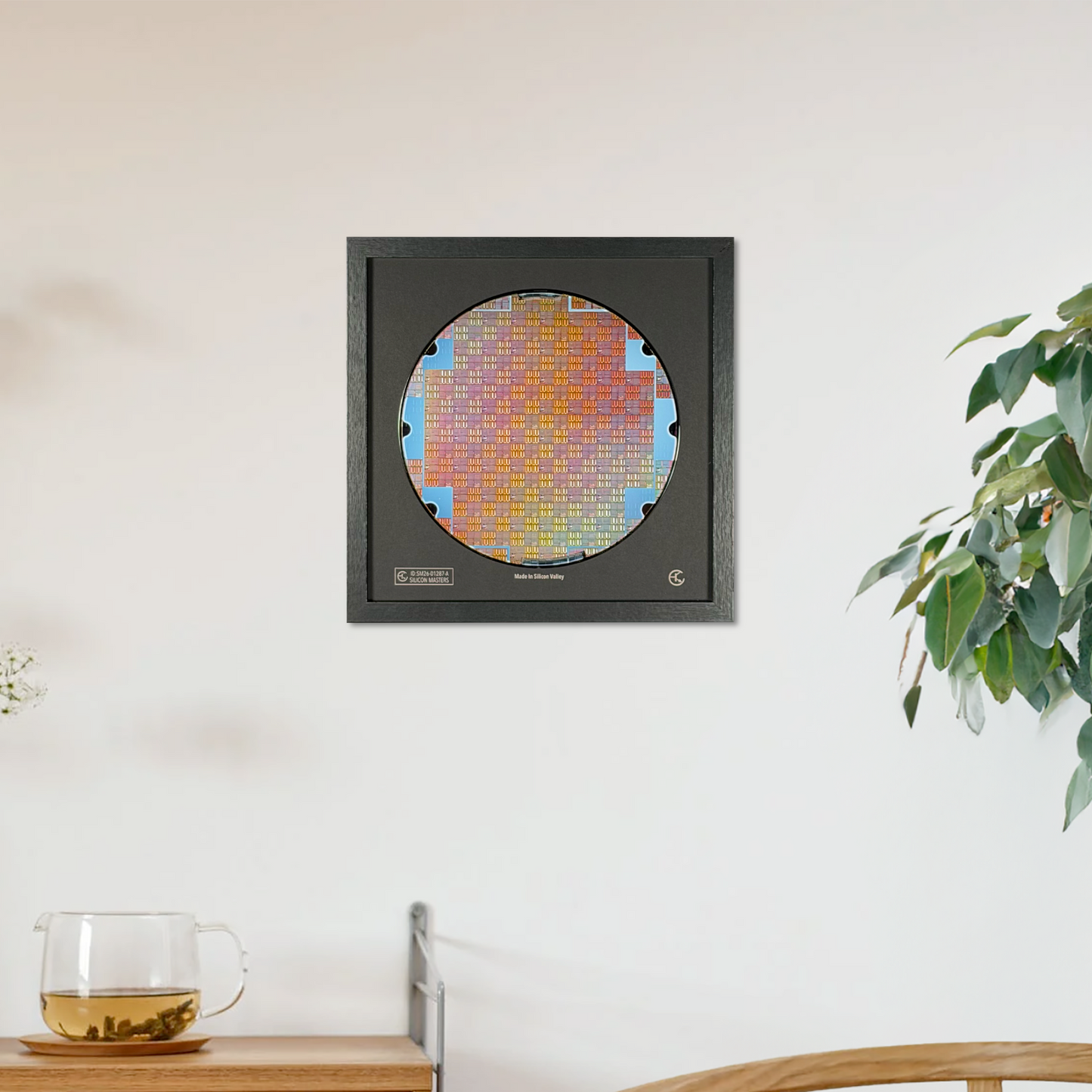 A square black-framed art print featuring a colorful circular pixelated design, displayed on a white wall in a modern room setting with a wooden cabinet, chair, and potted plant.