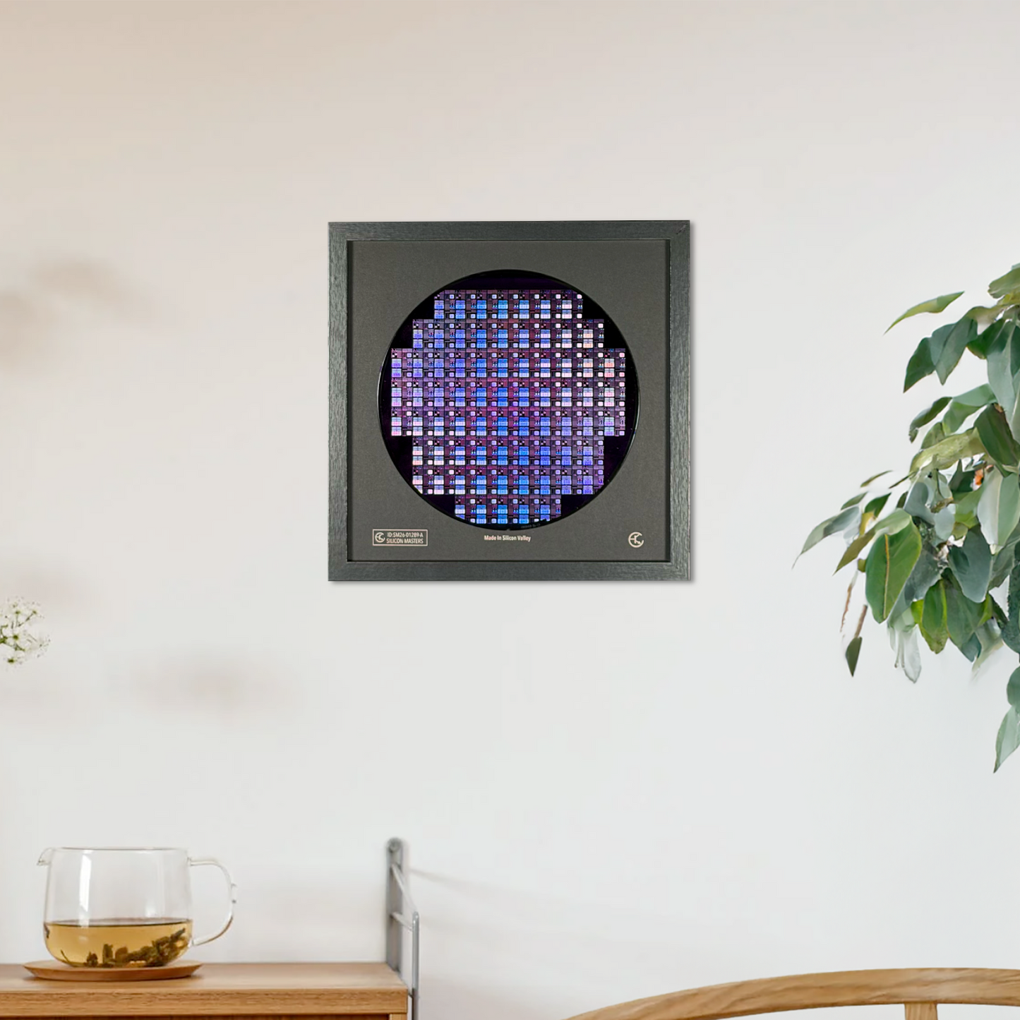 A square wall-mounted silicon wafer with a circular glowing blue-purple grid, shown hanging on a white wall in a styled interior with a wooden cabinet, chair, and potted plant.