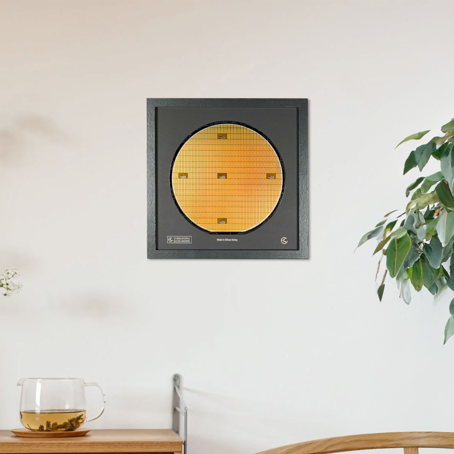 A square black wall-mounted display featuring a circular gold-colored textured panel, shown hanging on a white wall in a styled interior with a wooden cabinet, glass cup, and potted plant.
